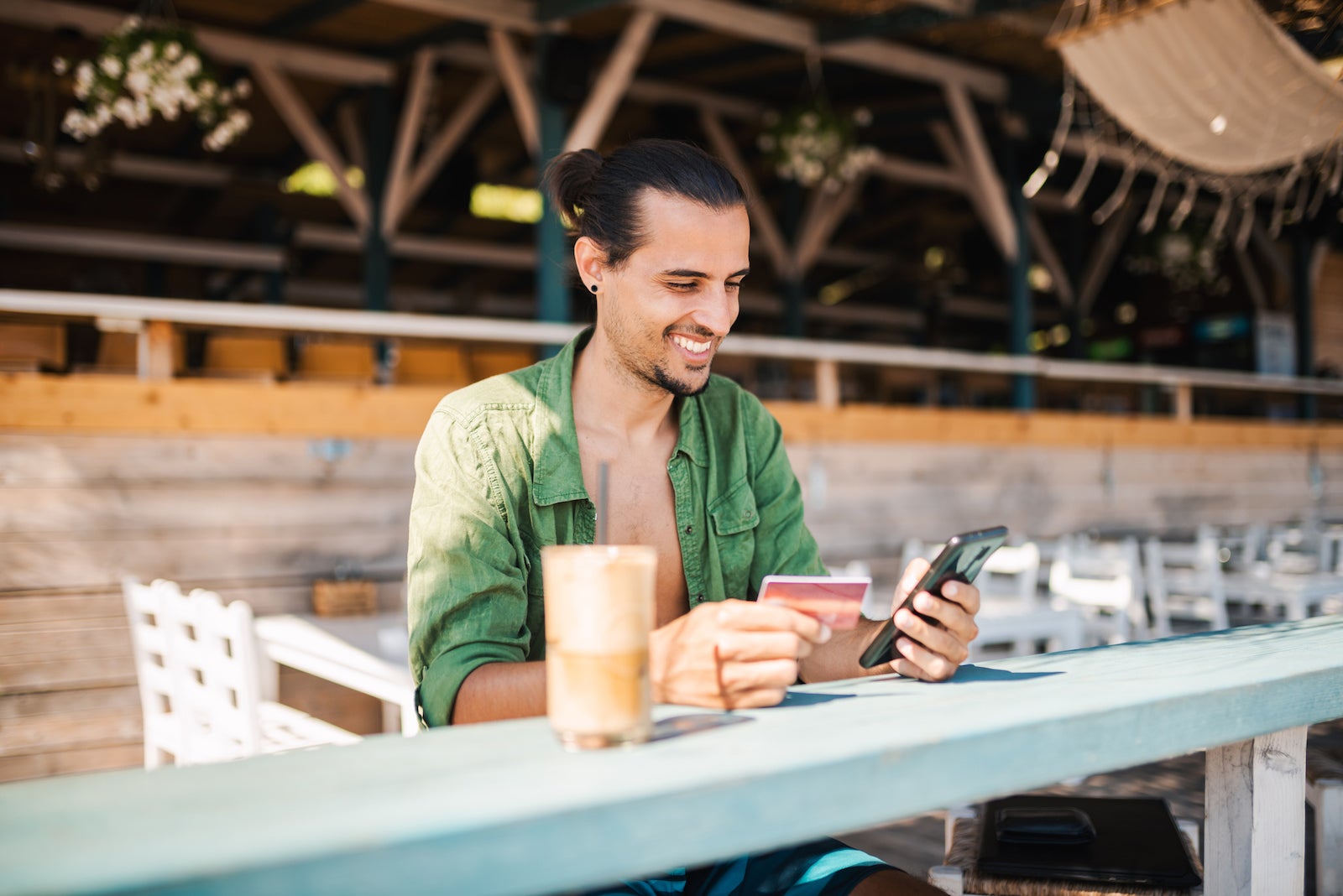 Young man on a beach bar with a cold Frappe coffee, shopping from his phone